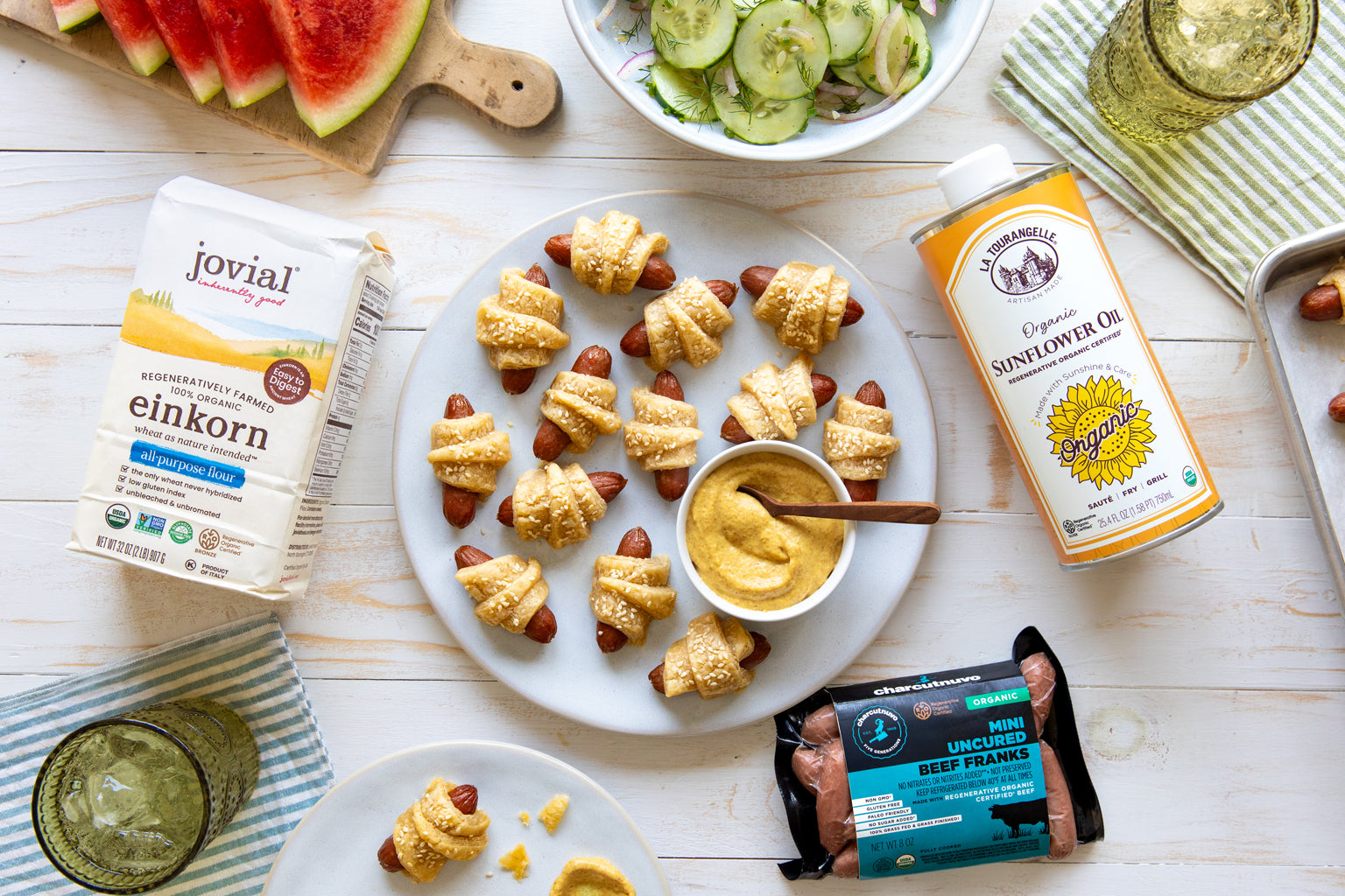 Overhead of pigs in a blanket on a plate with mustard, alongside Jovial einkorn flour and La Tourangelle sunflower oil