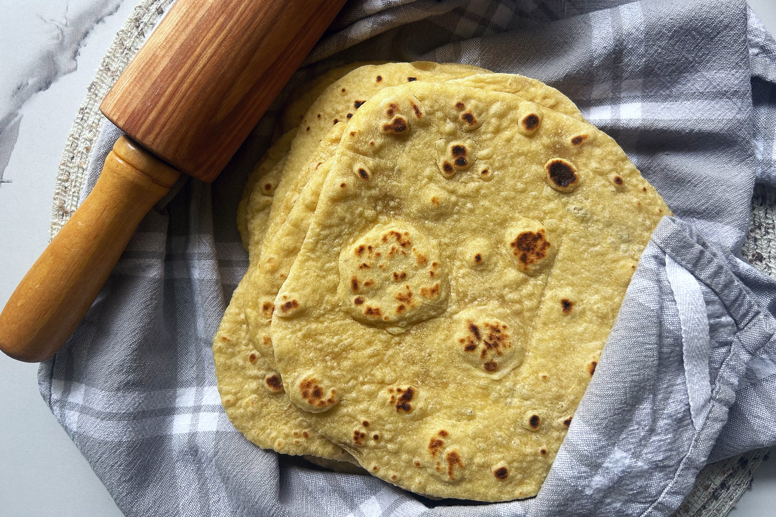Top-down view of two golden sourdough tortillas with charred bubbles on a gray checkered cloth beside a wooden rolling pin
