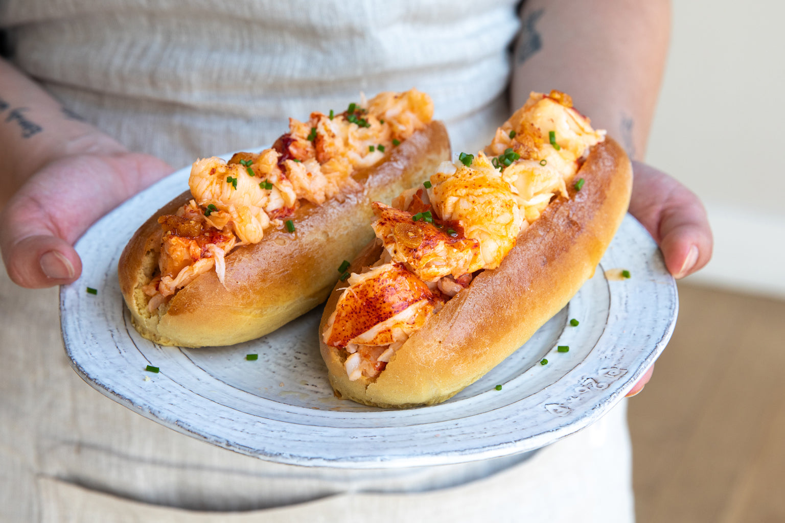 Two warm, buttery lobster rolls in toasted split-top buns garnished with chives on a ceramic plate held by a person