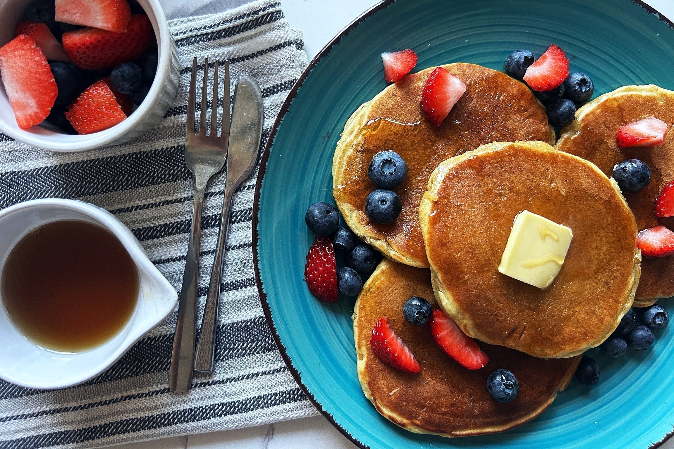 Three sourdough pancakes with butter and fresh strawberries and blueberries on a blue plate, syrup and forks on a striped napkin