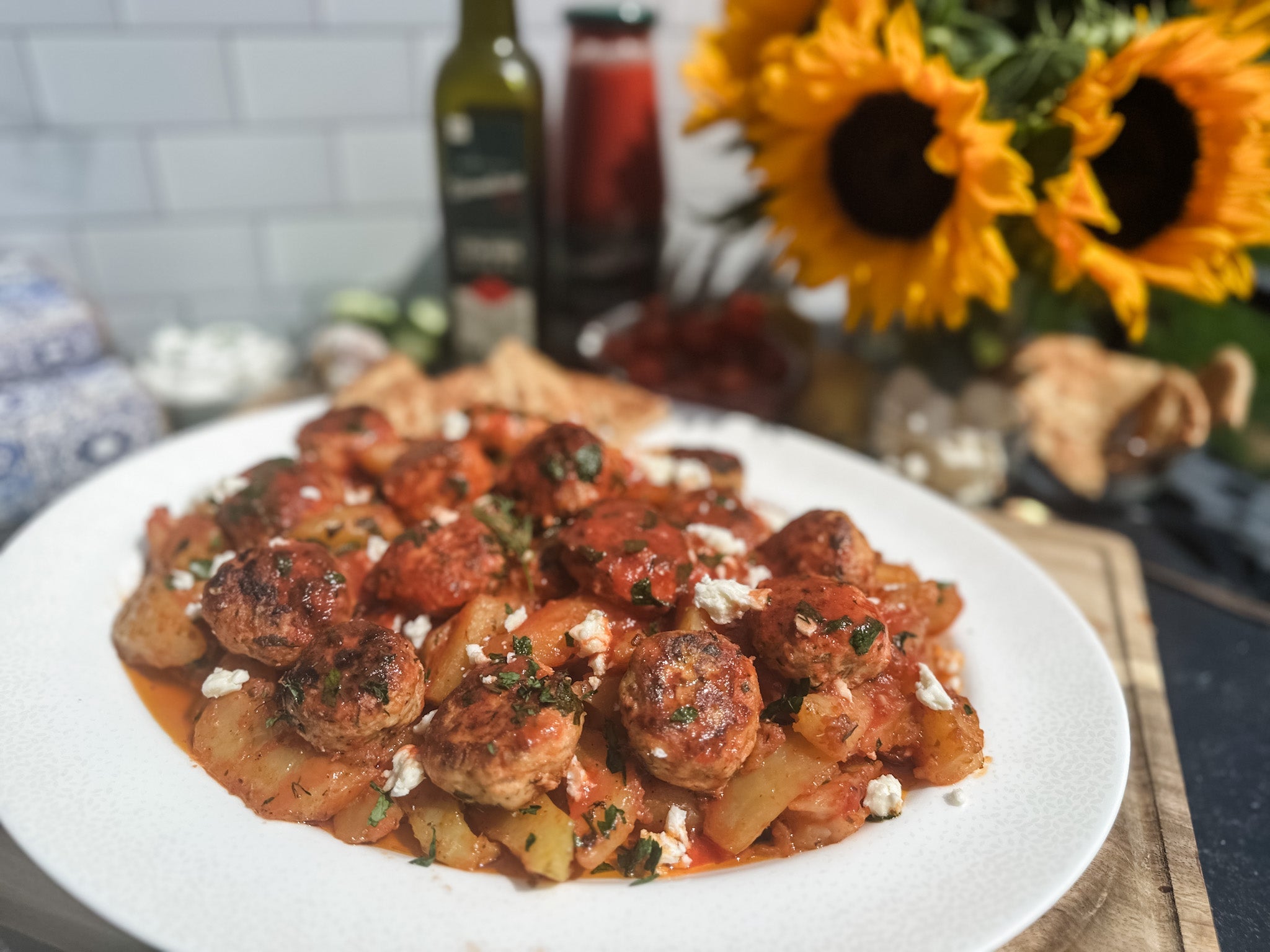 Greek chicken meatballs with roasted potatoes on a white plate, tomato sauce and feta, sunflowers blurred behind