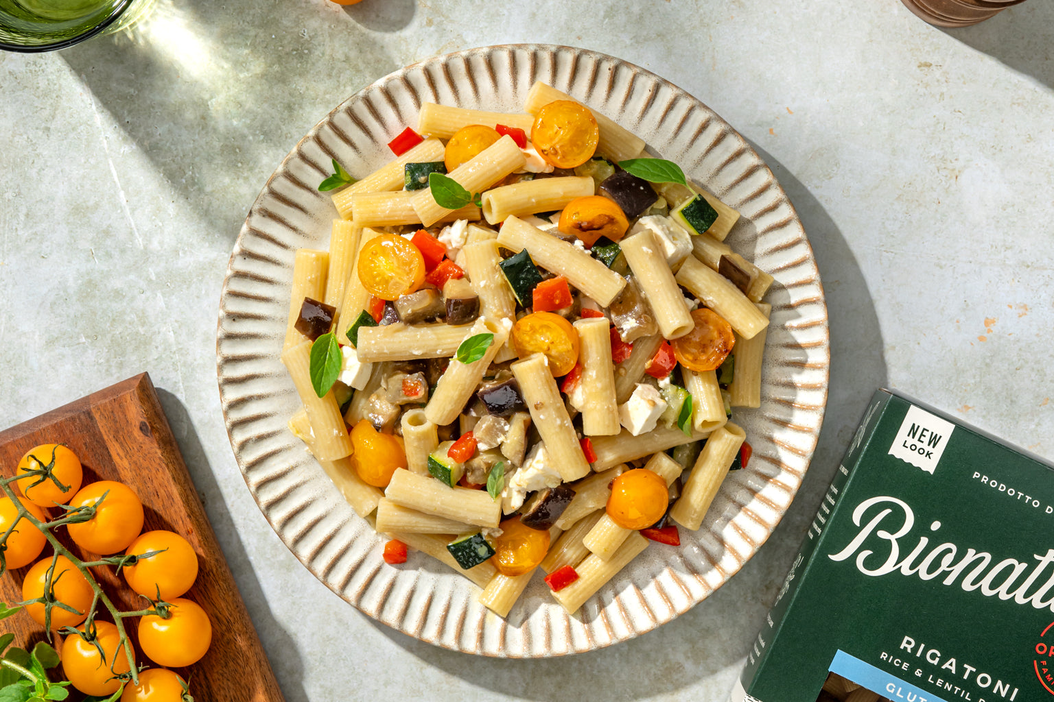Plate of rigatoni tossed with roasted eggplant, zucchini, yellow cherry tomatoes and crumbled feta next to a Bionaturae rigatoni box on a marble surface