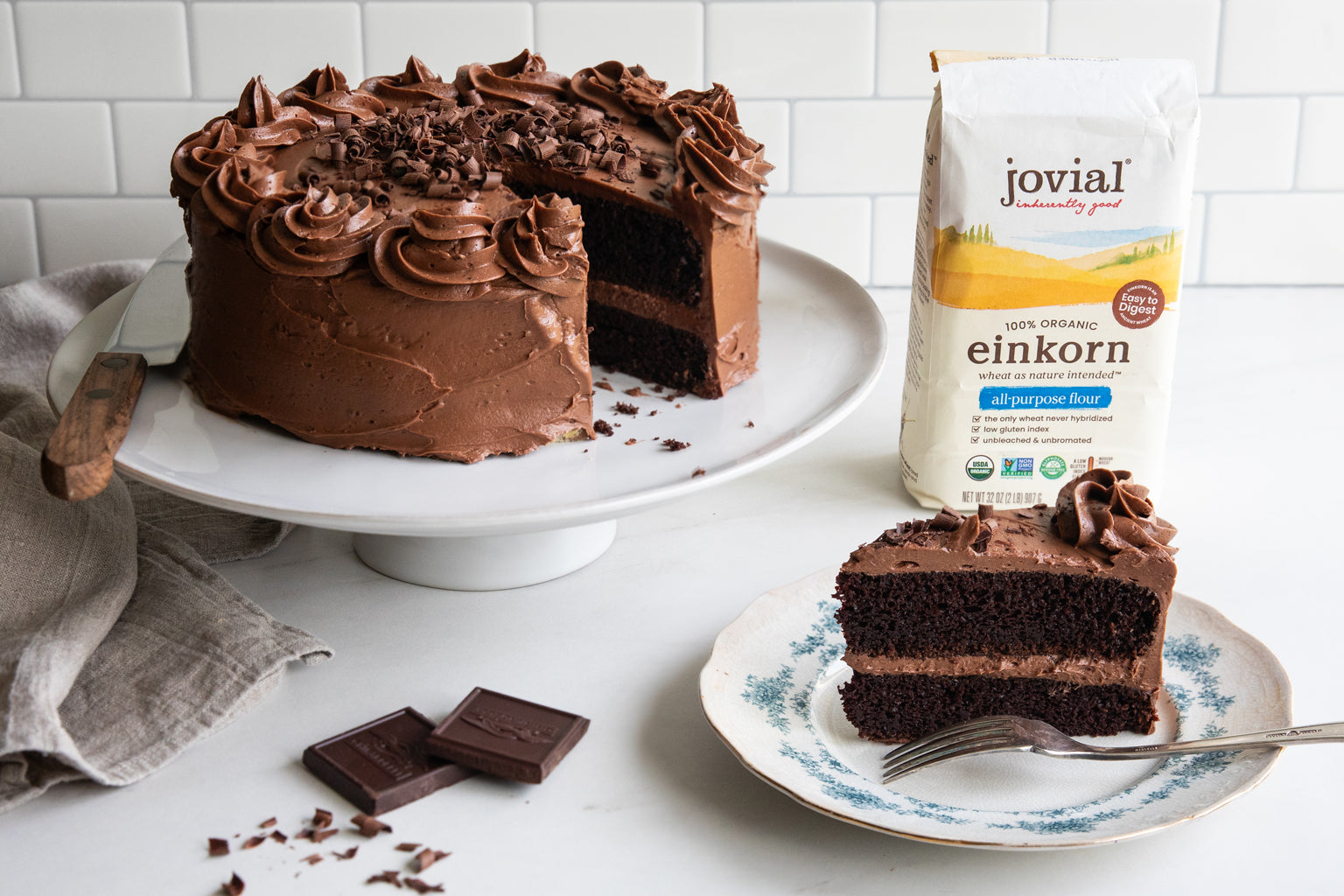 Chocolate layer cake sliced on a plate with chocolate frosting, cake on a pedestal and a bag of Jovial Einkorn flour in the background