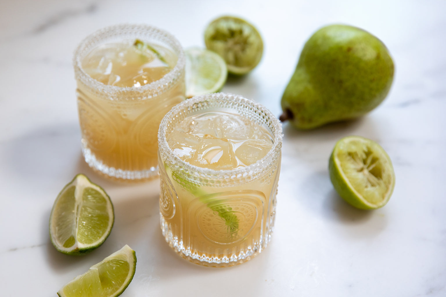 Two pear nectar margaritas on ice in cut-glass tumblers with salted rims, lime wedges and a pear on a marble surface