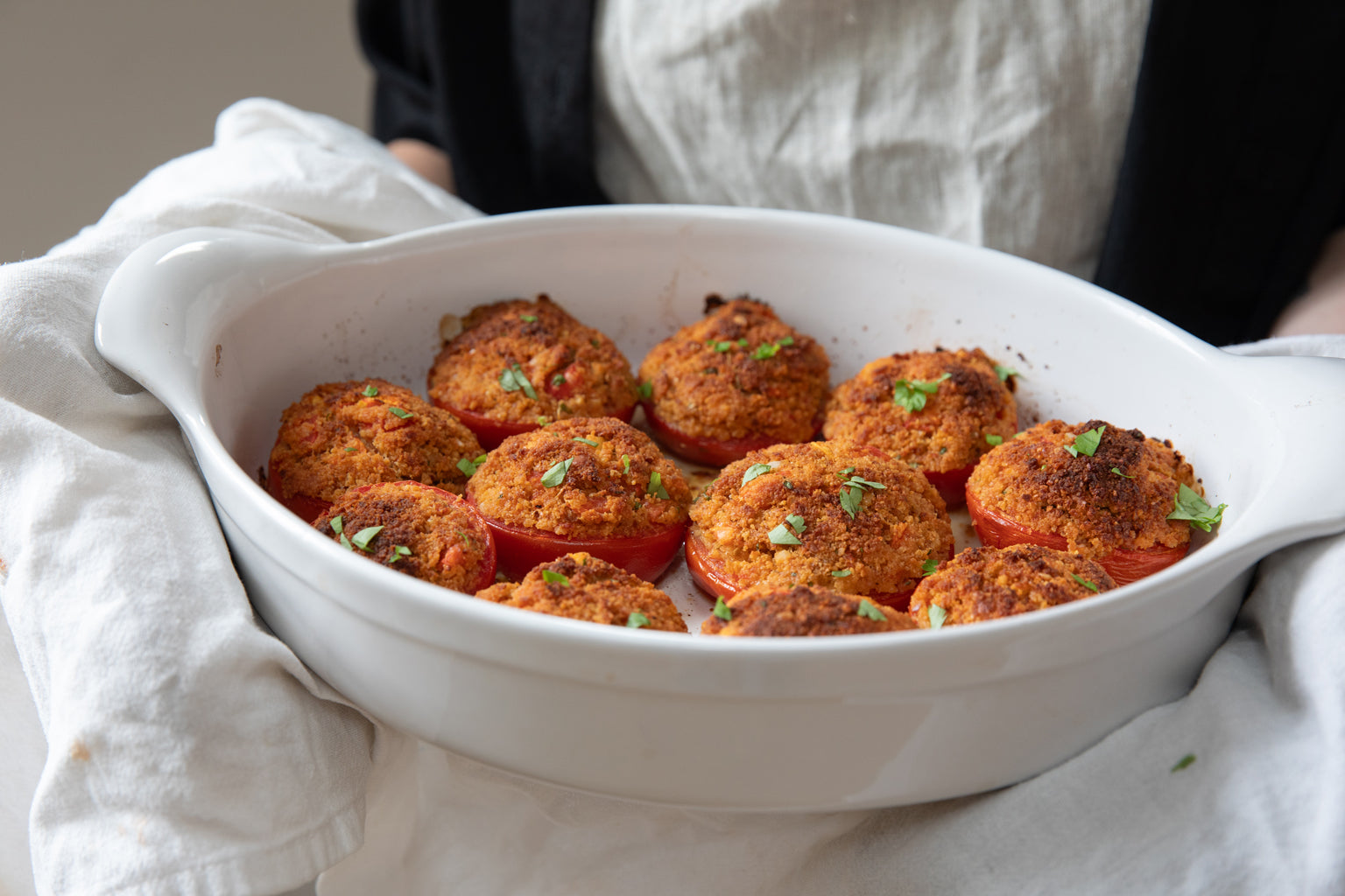 Baked tomato halves topped with golden einkorn breadcrumbs in a white oval baking dish held in a linen towel