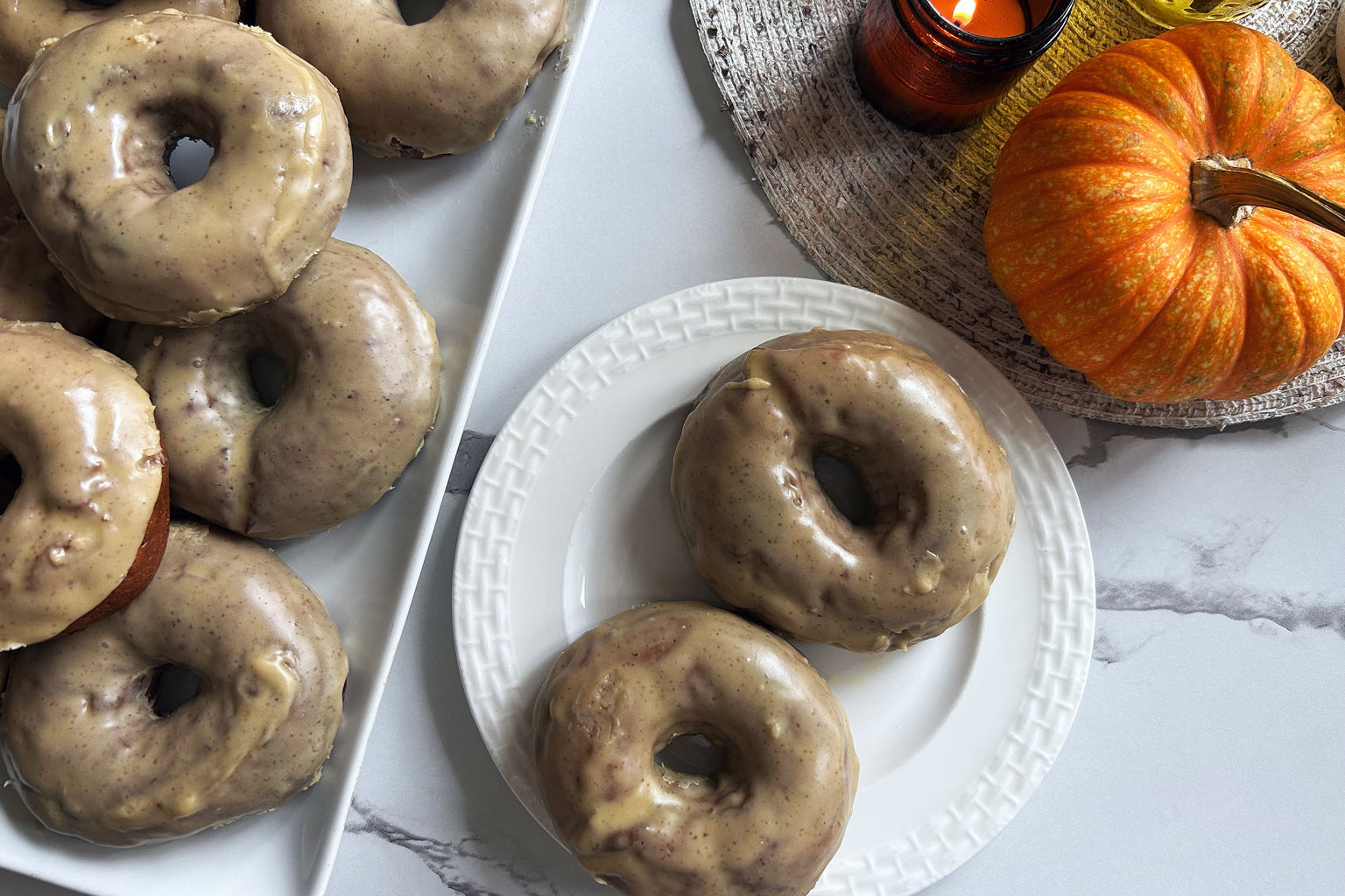 Overhead of sourdough pumpkin einkorn donuts showing speckled glaze on a white plate with a small pumpkin on marble