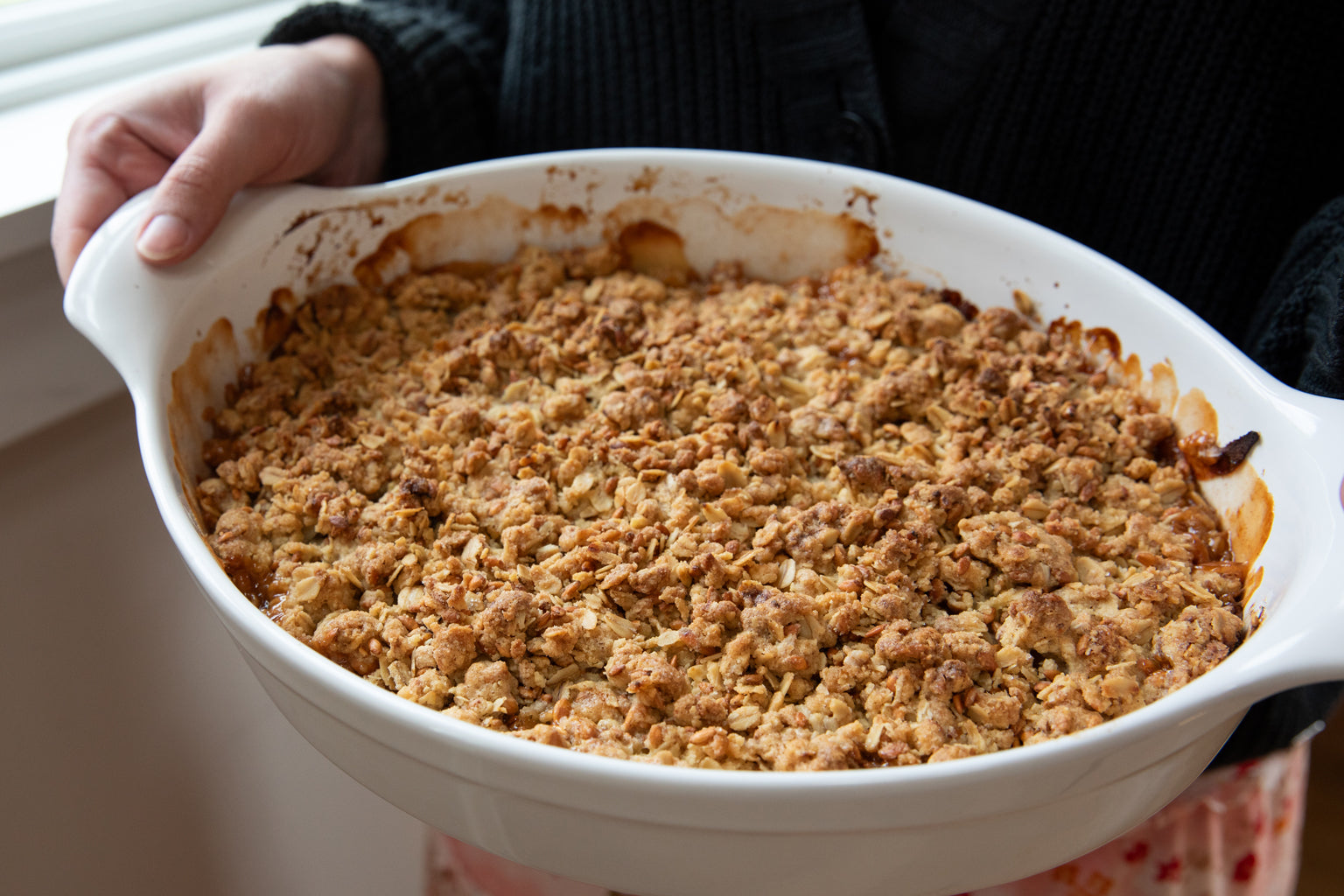Baked Einkorn apple crumble in a white oval baking dish being held by a person near a window