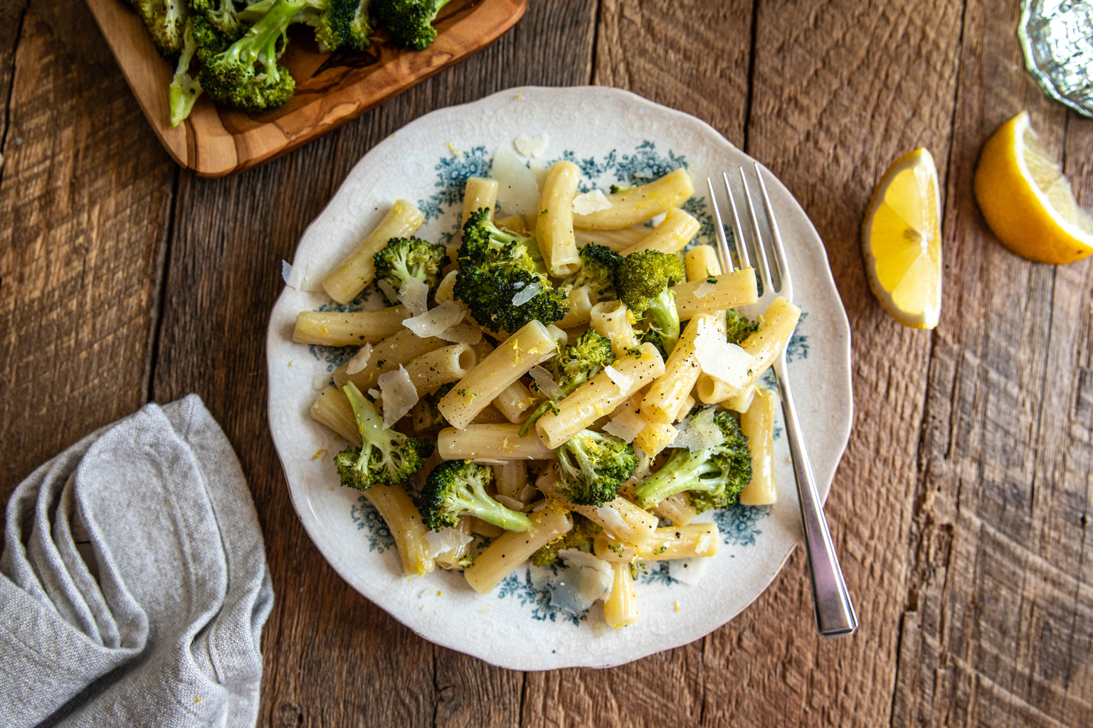 Rigatoni and roasted broccoli served on a floral plate on a rustic wooden table with a fork and lemon wedges