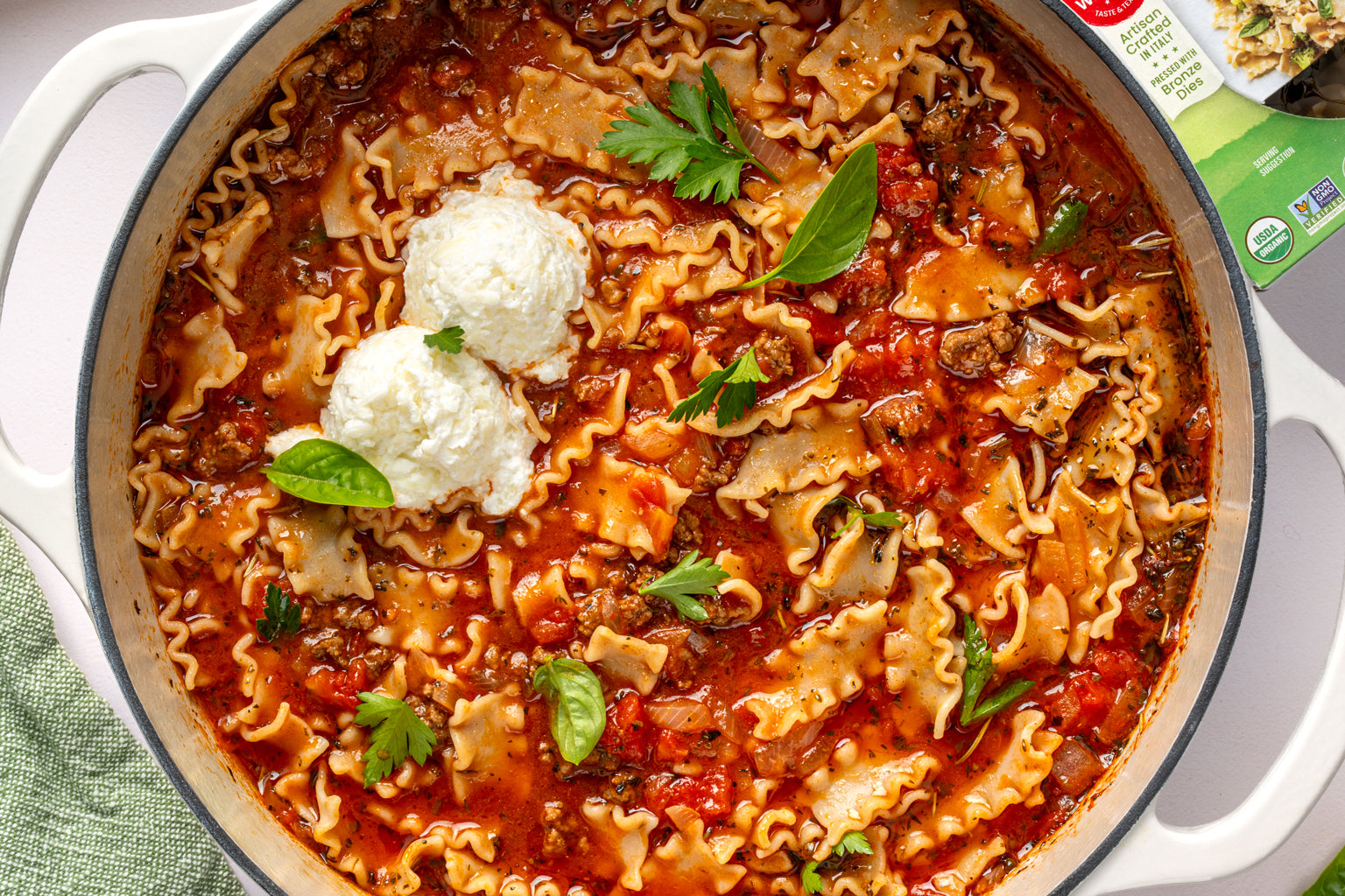 Lasagna soup in a white pot with gluten free mafalda pasta, tomato-meat broth, fresh basil, and two dollops of ricotta