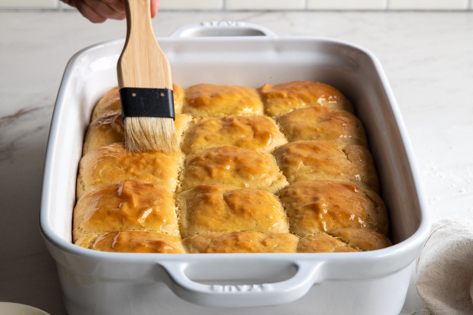 Einkorn dinner rolls being brushed with melted butter in a white Staub baking dish on a kitchen counter