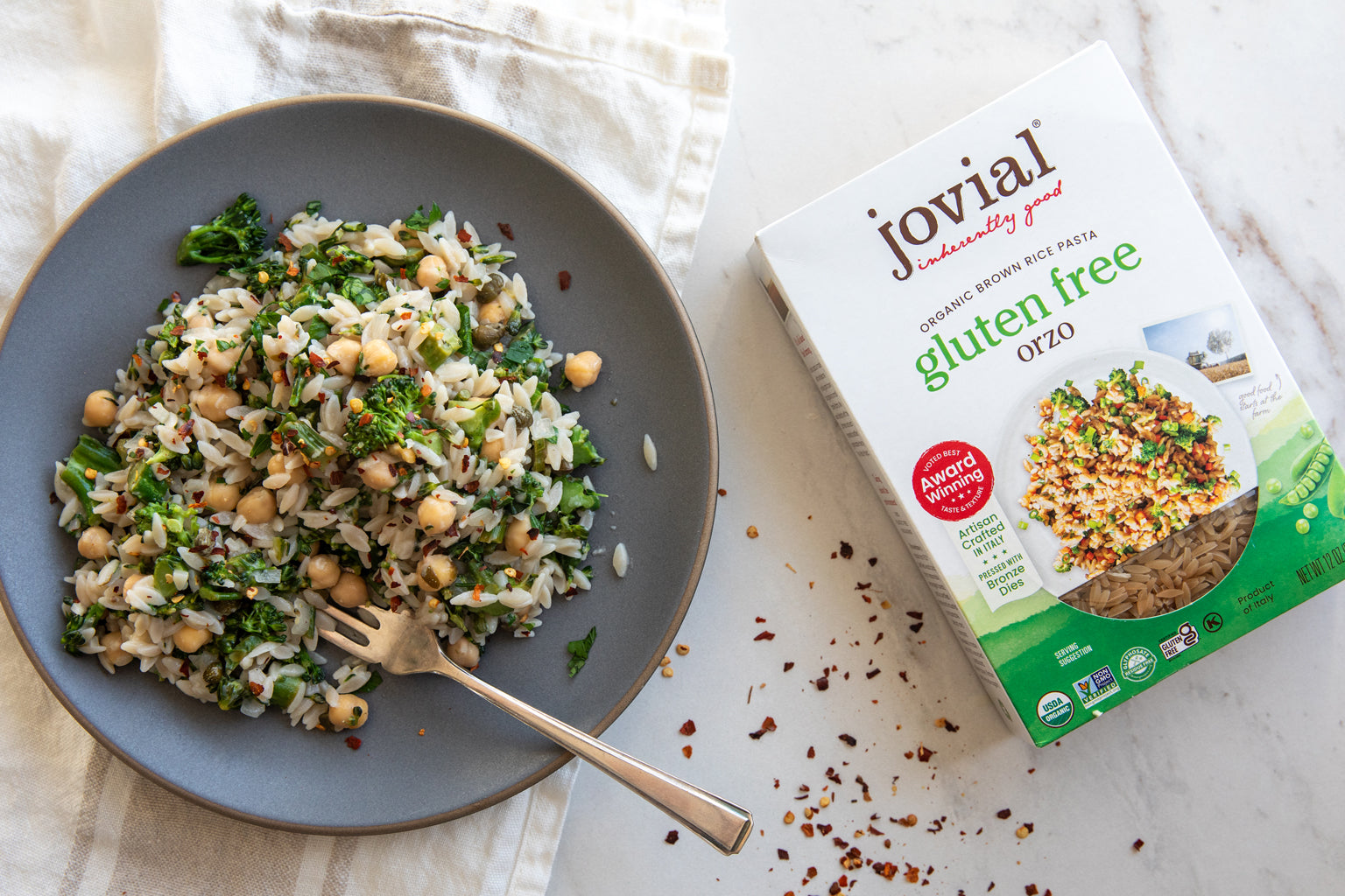 Broccolini orzo with chickpeas on a gray plate with a fork, beside a Jovial gluten free orzo box on a light marble surface