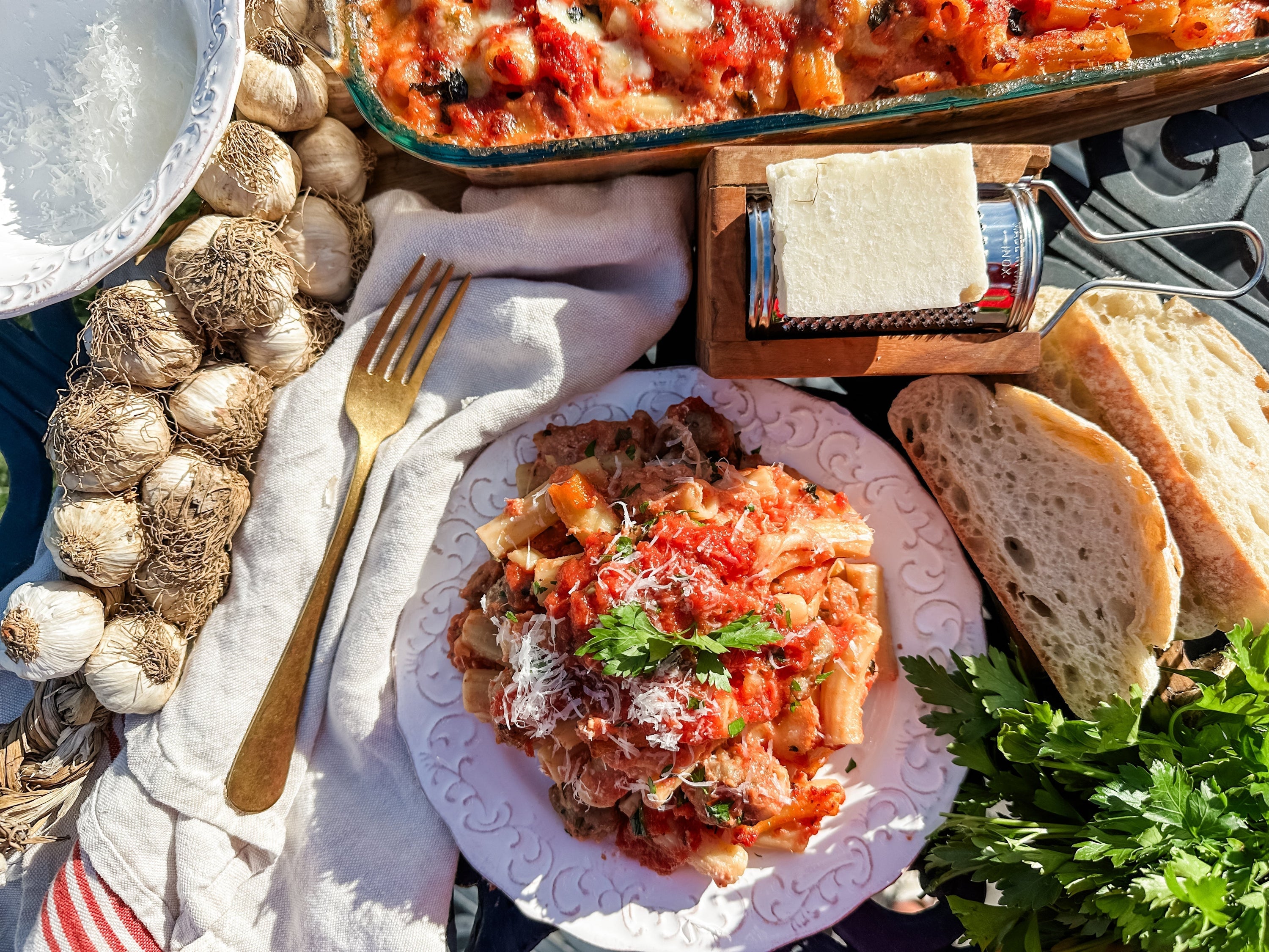 Baked rigatoni with mini sausage meatballs on a white plate, grated cheese and parsley, with crusty bread and garlic nearby