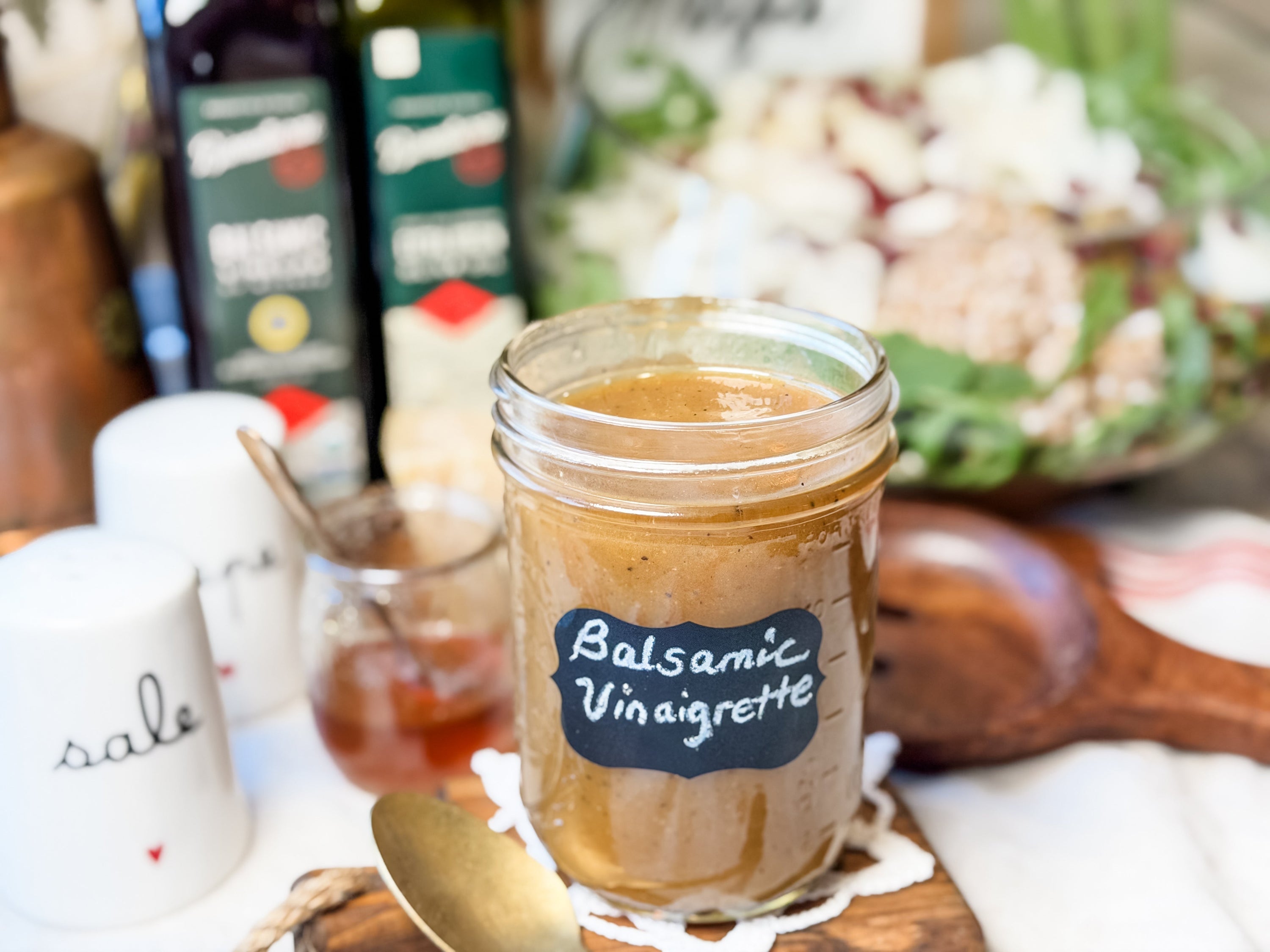 Mason jar labeled Balsamic Vinaigrette on a wooden board with a spoon, salad and olive oil bottles blurred in the background