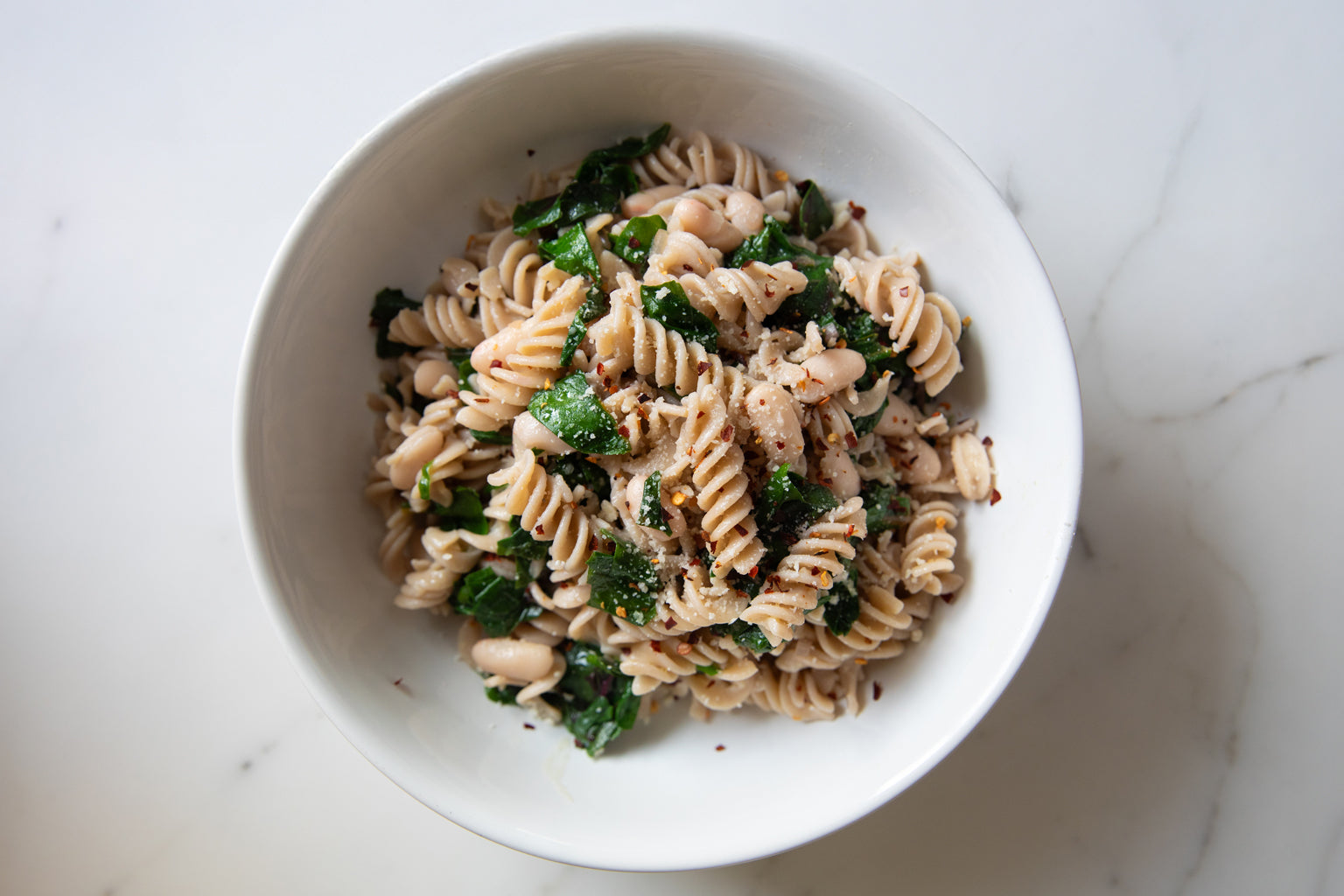 Bowl of gluten-free fusilli tossed with Swiss chard and cannellini beans on a marble countertop