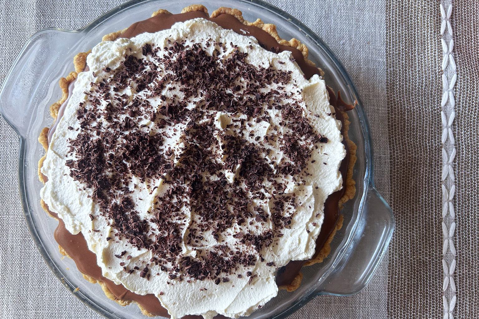 Top view of French silk pie with whipped cream and chocolate shavings in a glass pie dish on a beige linen tablecloth