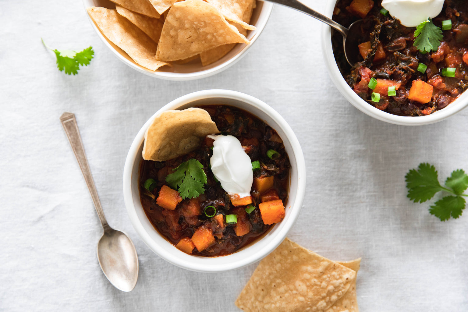 Overhead view of two bowls of vegetarian chili with butternut squash and kale, topped with sour cream, cilantro, green onions, and tortilla chips