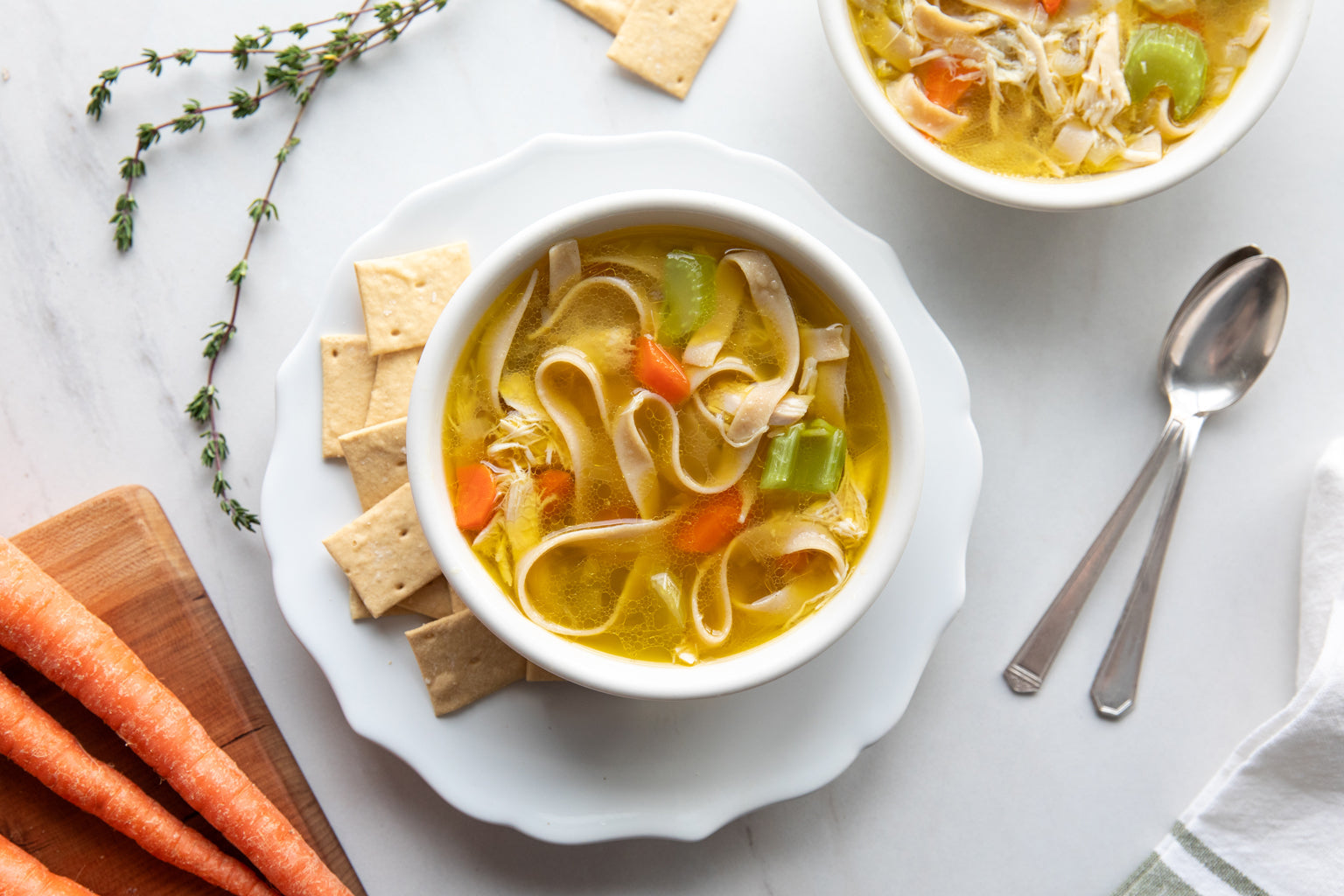 Two bowls of chicken noodle soup with brown rice egg tagliatelle, carrots and celery on marble table with crackers and spoons