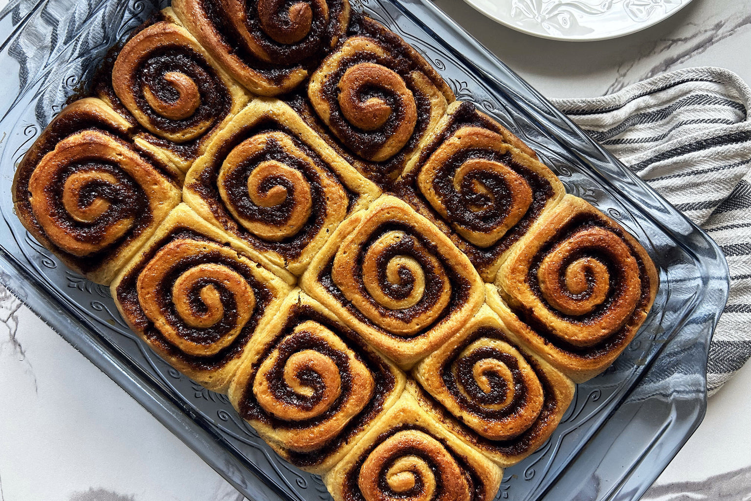 Sourdough cinnamon rolls cooling in a glass baking dish on a marble countertop beside a striped kitchen towel