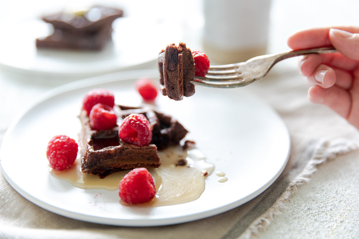 Dark chocolate Einkorn waffle with raspberries and syrup, fork lifting a bite on a white plate