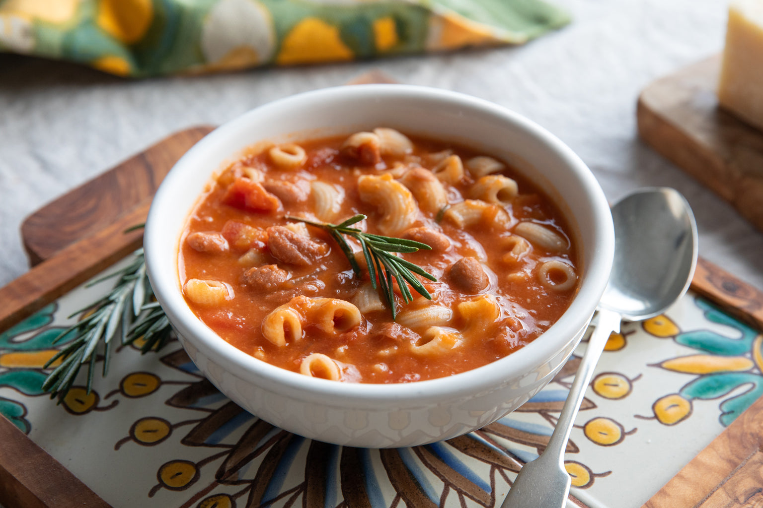 Bowl of pasta e fagioli soup garnished with a rosemary sprig, served on a patterned wooden tray with a spoon