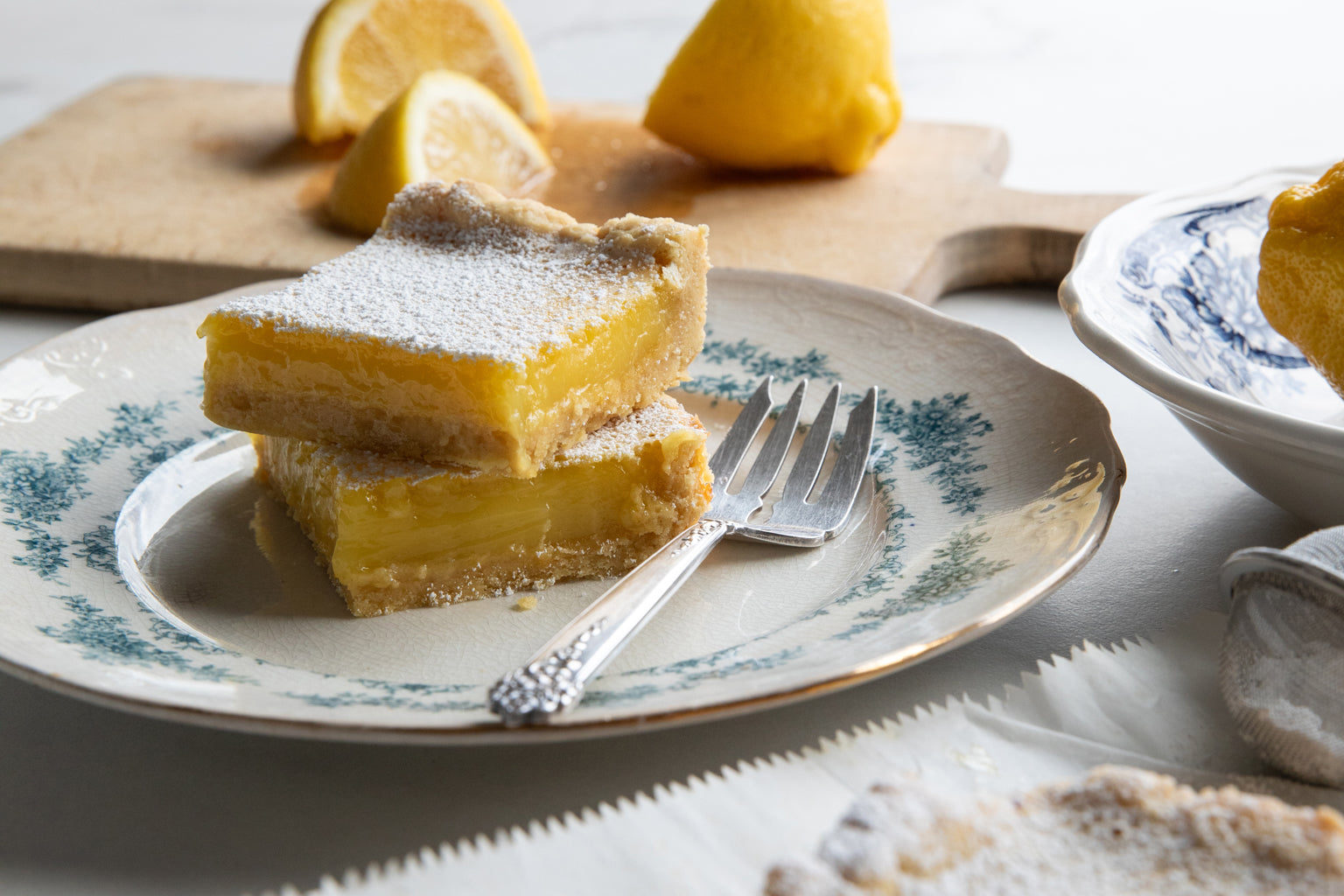 Two stacked Einkorn lemon bars dusted with powdered sugar on a vintage plate with a fork and lemon wedges in the background