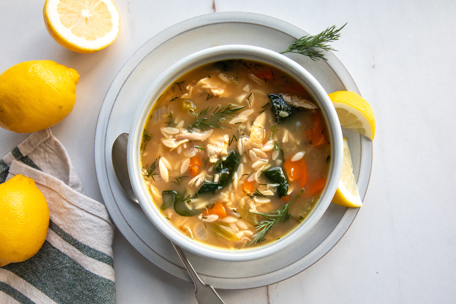Single bowl of lemon orzo chicken soup with spinach and dill, served on a gray plate with lemon wedges and whole lemons nearby