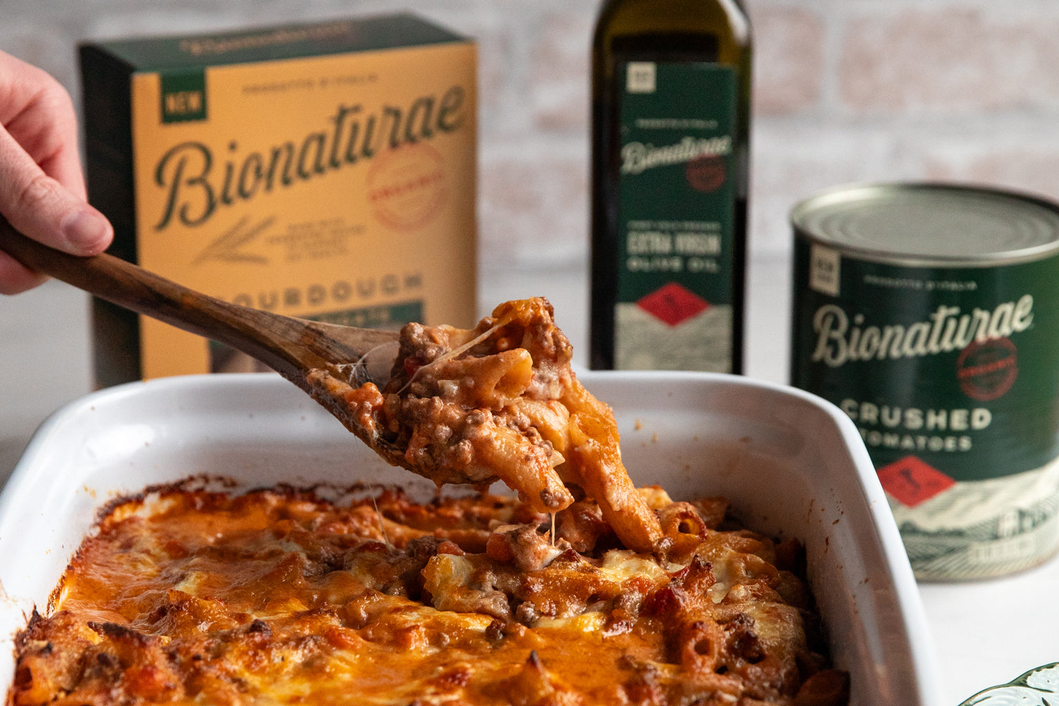 Baked sourdough penne al forno being lifted from a casserole with melted cheese, Bionaturae products in the background