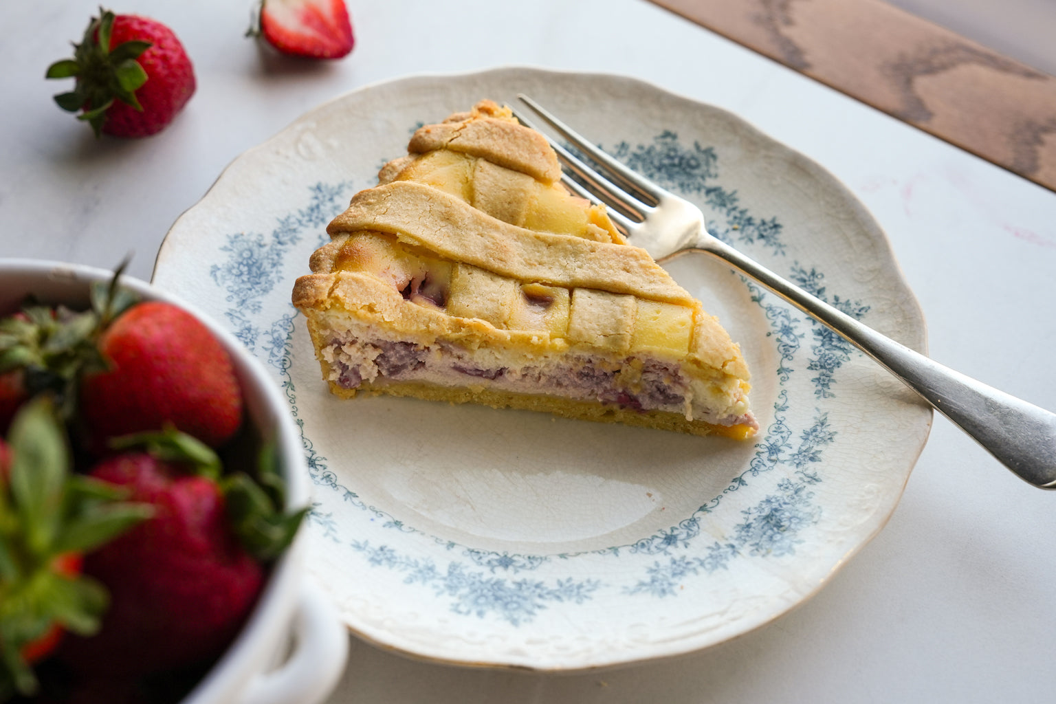Angled view of ricotta and strawberry einkorn crostata slice with lattice crust and creamy filling on a floral plate