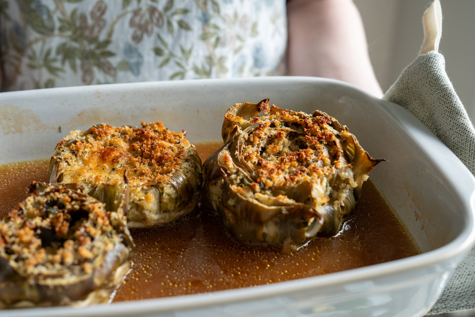 Three baked stuffed artichokes in a shallow white baking dish with golden breadcrumb topping and cooking broth