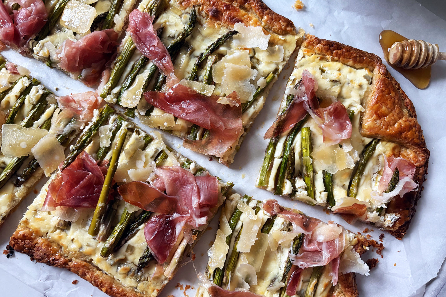 Sliced asparagus tart on parchment with sourdough crust, shaved Parmesan, prosciutto, and honey dipper