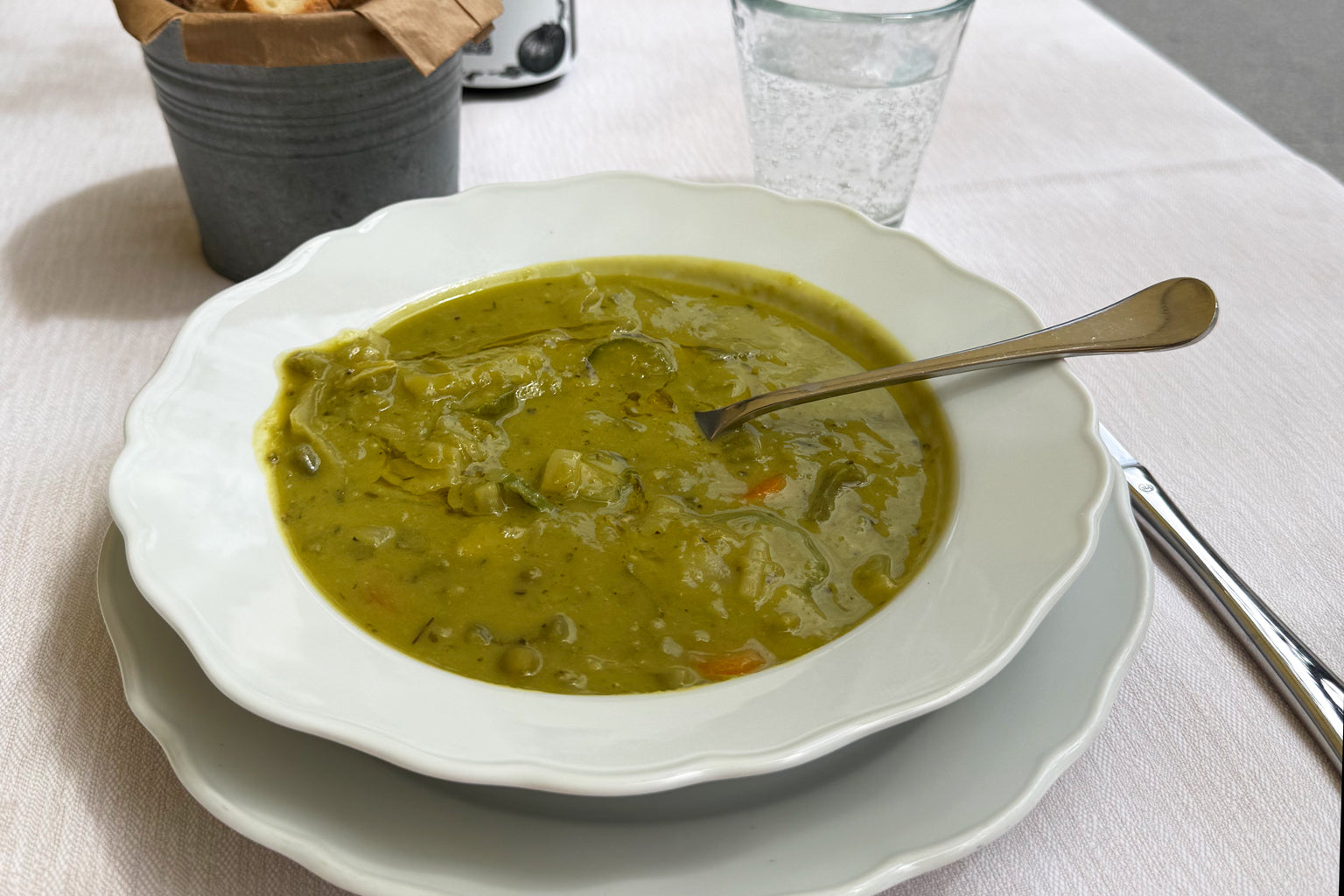 Bowl of Tuscan pea soup in a white scalloped bowl with a spoon, on a white tablecloth with a glass of water nearby