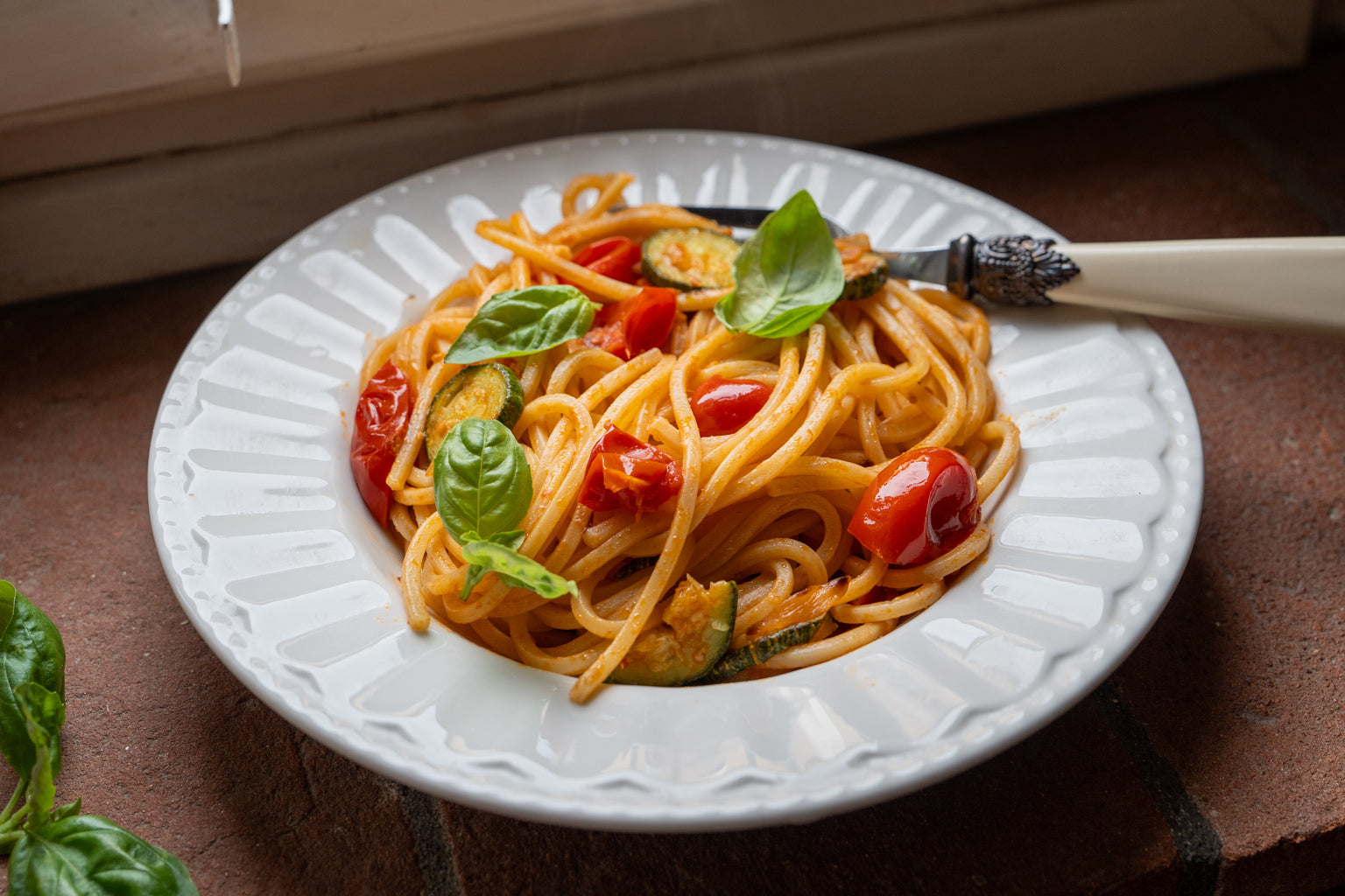 Plate of spaghetti with zucchini, cherry tomatoes, and fresh basil on a white ceramic plate by a windowsill