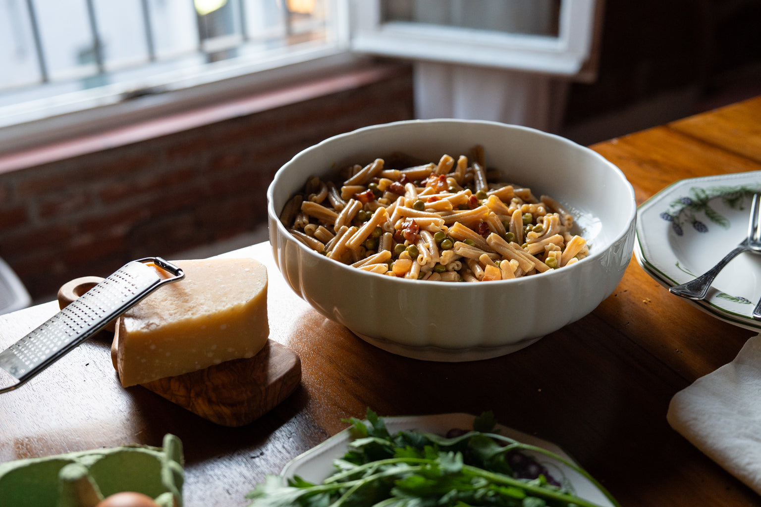 Bowl of caserecce carbonara with peas on a wooden table by a window, with a wedge of Parmesan and fresh parsley