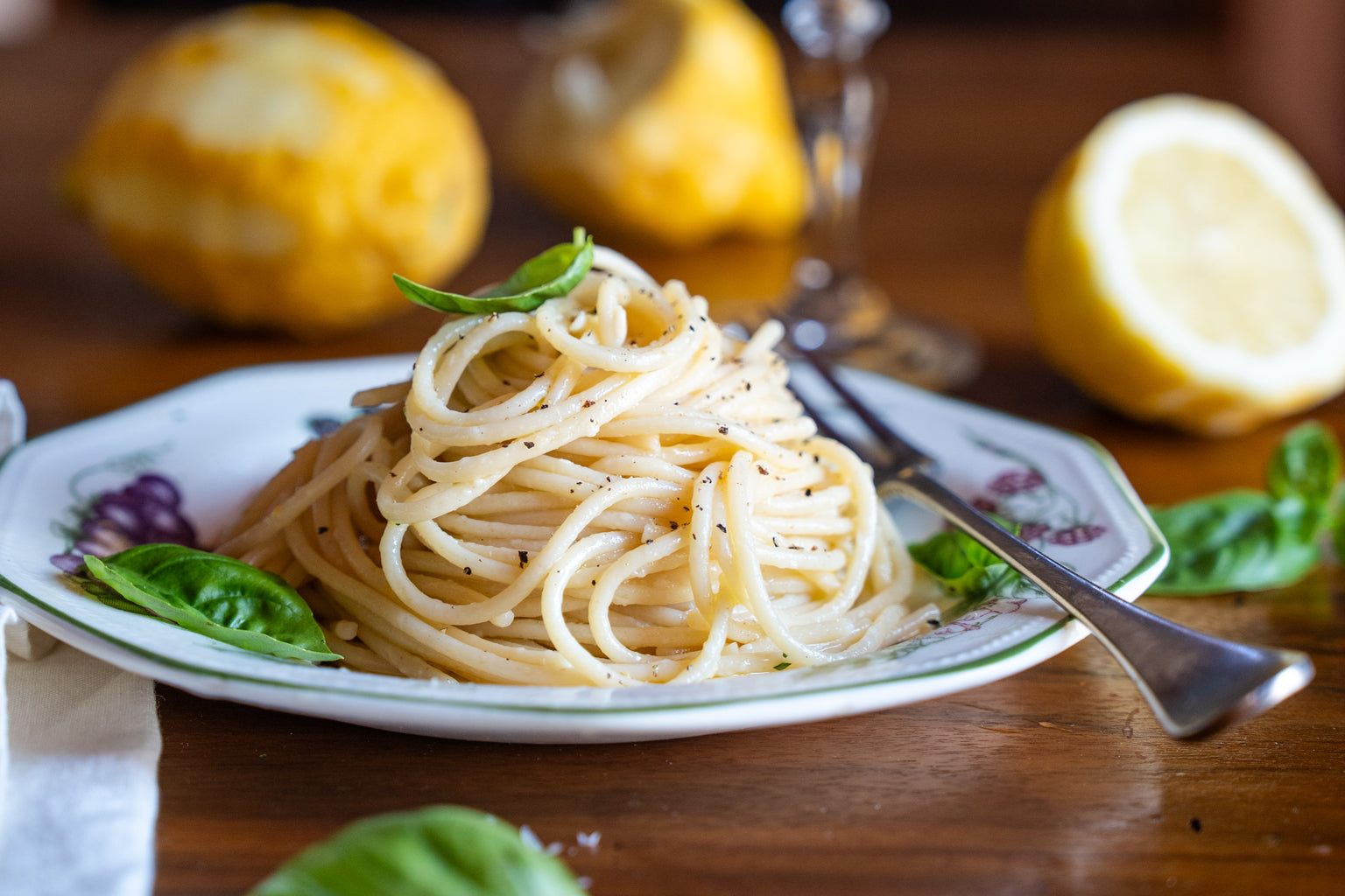 Plate of Spaghetti al limone on a wooden table with basil leaves, cracked black pepper, lemon halves, and a fork