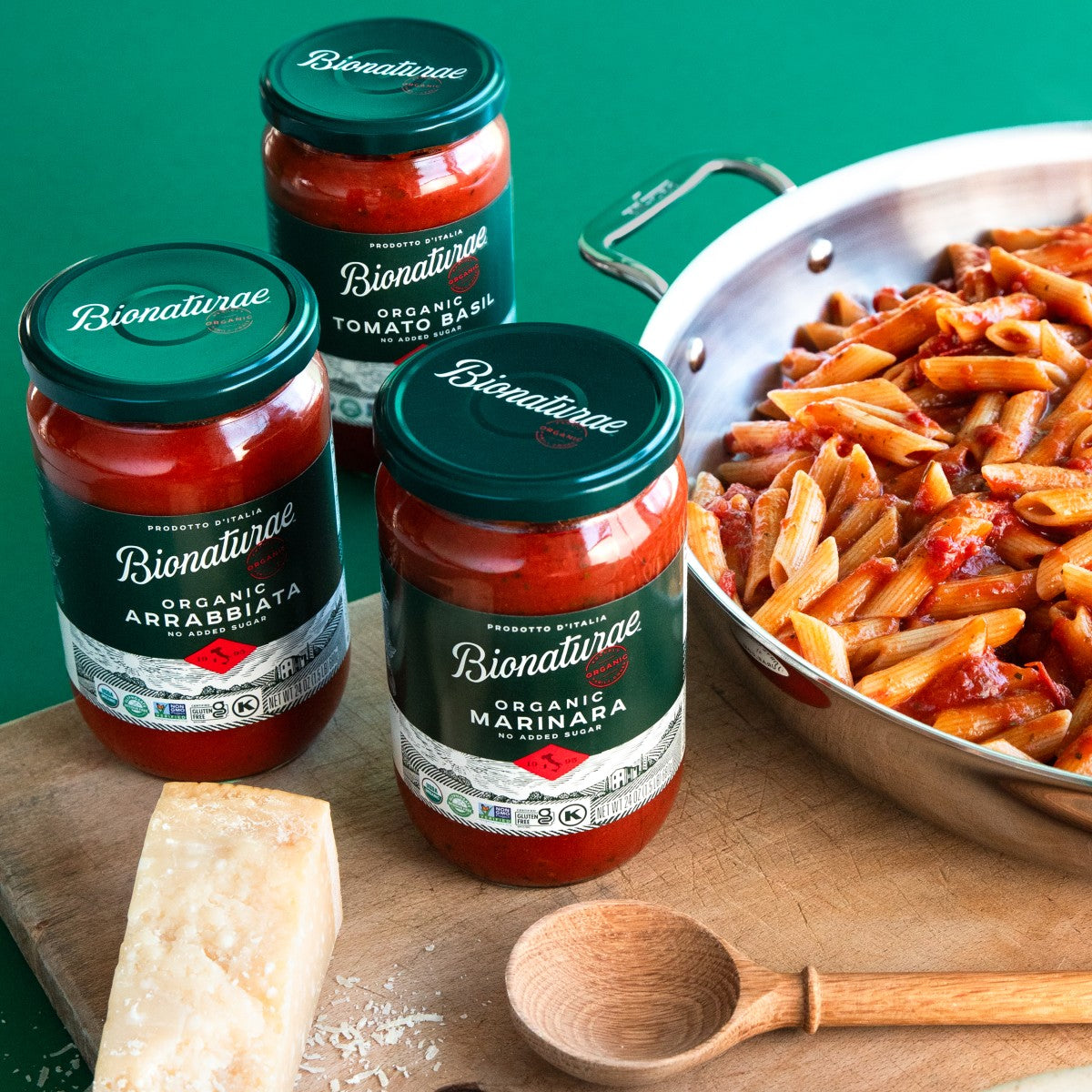 Three jars of Bionaturae organic pasta sauces on a wooden board with a skillet of penne and a wedge of Parmesan