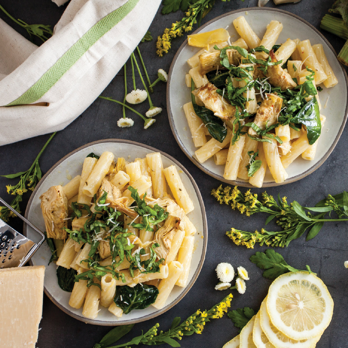 Two plates of gluten-free rigatoni with artichokes, wilted greens, and grated cheese on a dark table