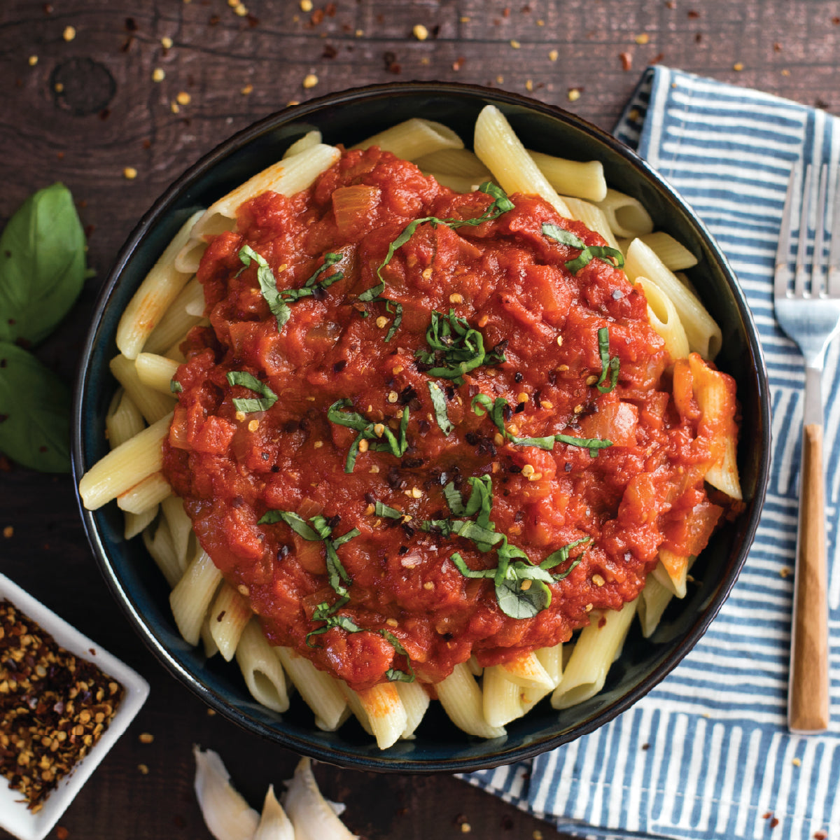 Bowl of gluten-free penne topped with chunky tomato sauce, basil, and red pepper flakes on a wooden table