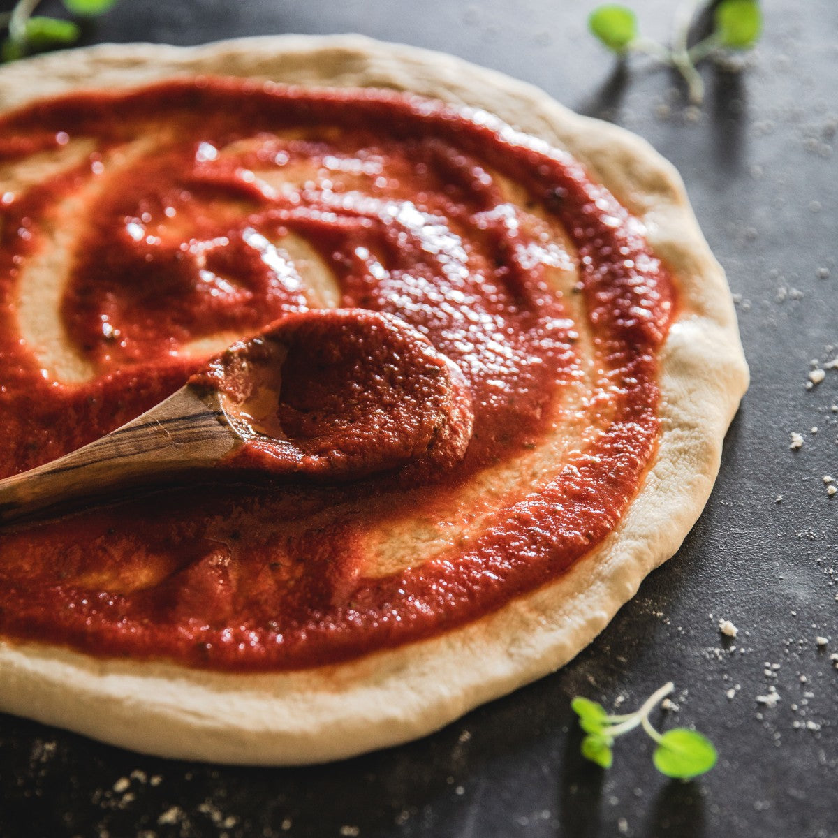 Wooden spoon spreading organic tomato sauce over pizza dough on a dark countertop