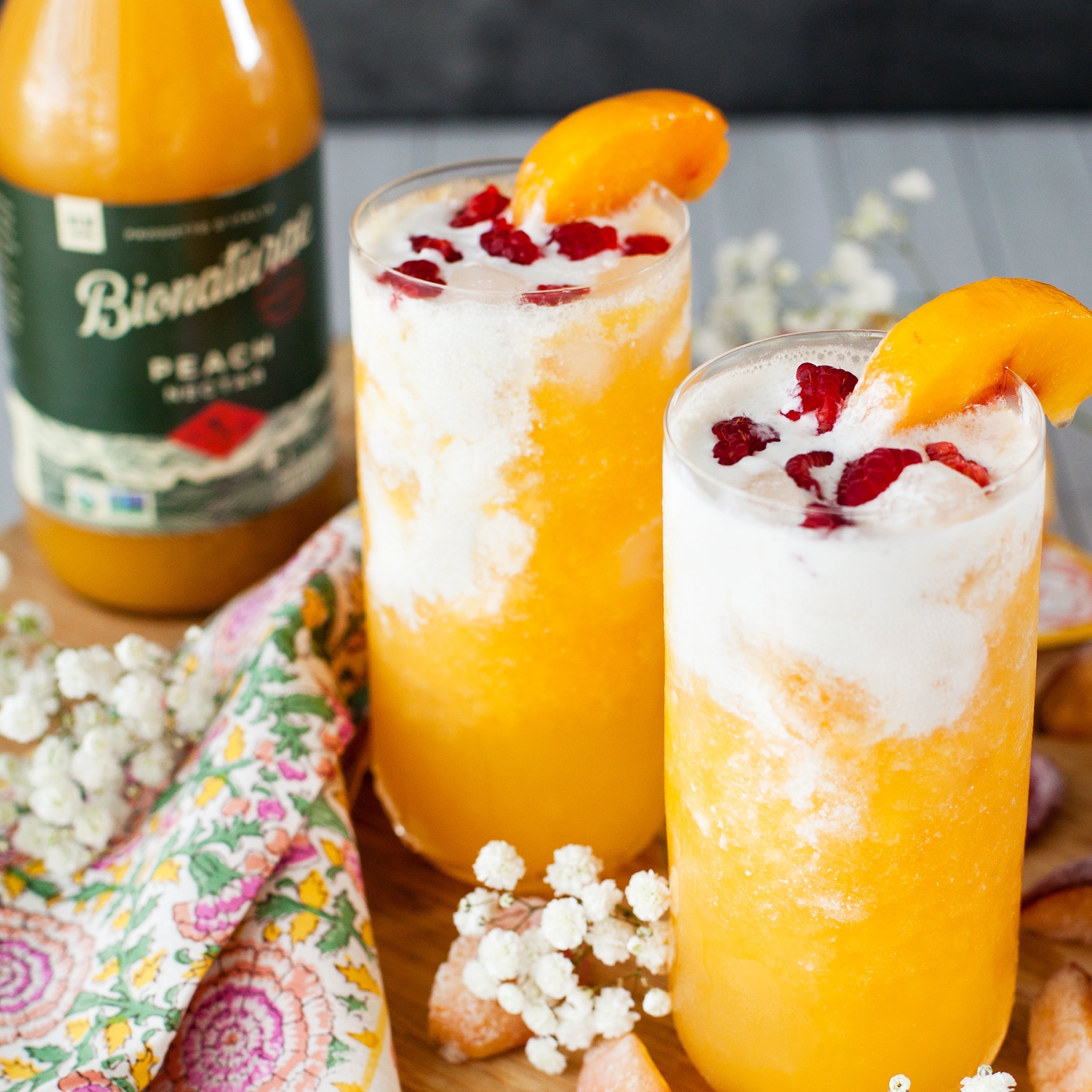 Two peach nectar floats topped with cream and raspberries, with a Bionaturae peach nectar bottle and floral napkin