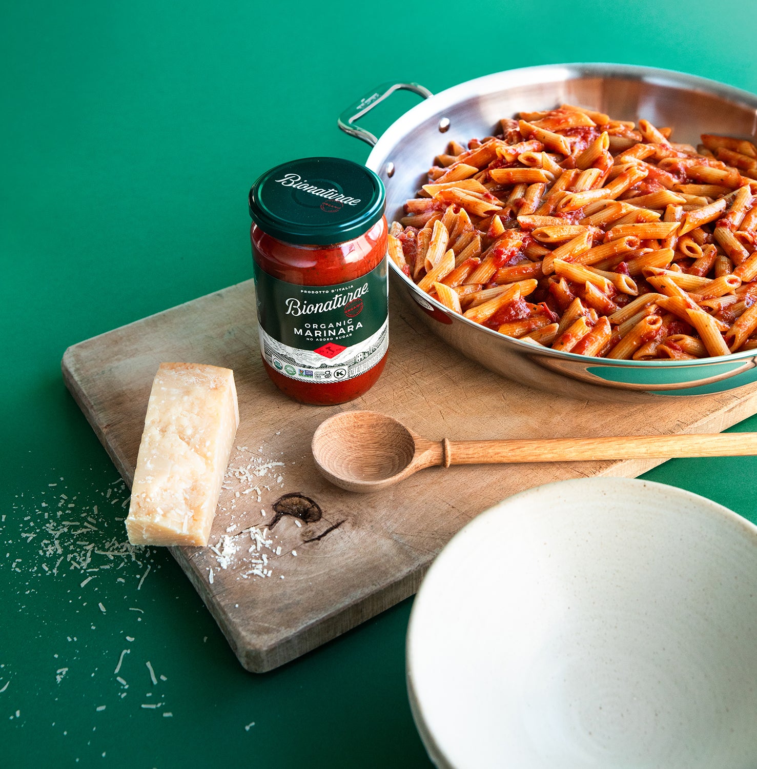 Jar of Bionaturae organic marinara sauce beside a bowl of penne with a wedge of parmesan and a wooden spoon on a green background