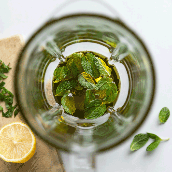 Top-down view of mint, honey and lemon dressing ingredients in a blender with olive oil, fresh mint leaves, and a lemon half on a cutting board