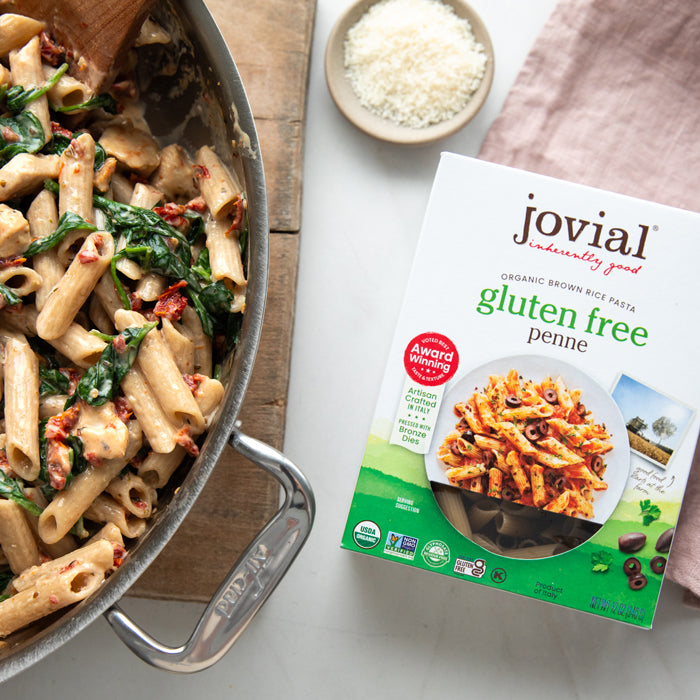 Skillet of creamy penne with spinach and sun-dried tomatoes beside a Jovial organic brown rice gluten-free penne box