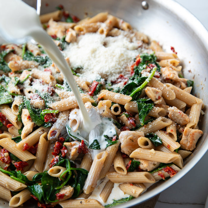 Skillet of chicken and Gluten Free penne with spinach, sun-dried tomatoes, cheese and cream being poured