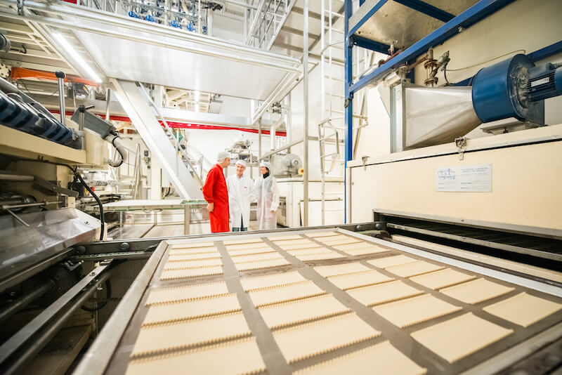 Three workers inspecting trays of rectangular pasta sheets on a production line inside an artisan pasta factory