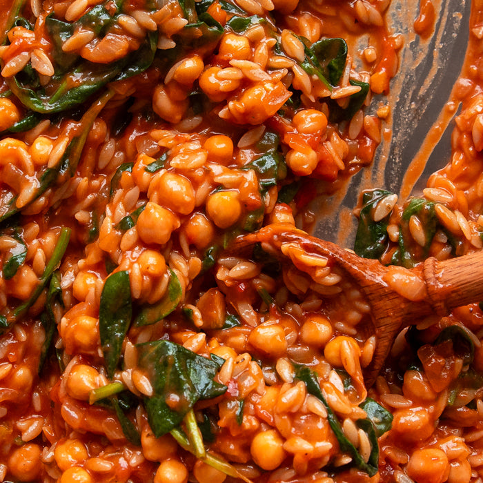 Close-up of saucy tomato chickpeas and brown rice orzo showing tender chickpeas, wilted spinach, and a wooden spoon in pan