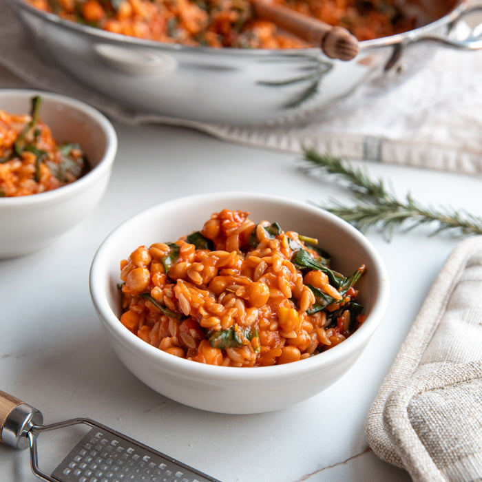Close up of Organic Gluten Free Brown Rice Orzo in a bowl showcasing saucy tomato, chickpeas, and herbs on a light tabletop
