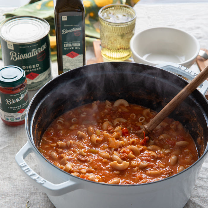 Dutch oven of steaming pasta e fagioli with tomatoes and a wooden spoon, bowl and glass in the background
