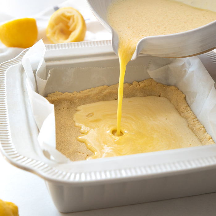 Bowl pouring lemon filling into an einkorn shortbread crust in a parchment-lined 8x8-inch baking pan