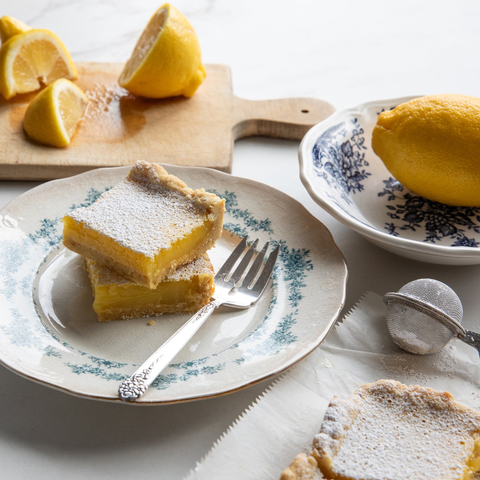 Two stacked lemon bar squares made with einkorn, dusted with powdered sugar, on a vintage floral plate with a fork and lemons
