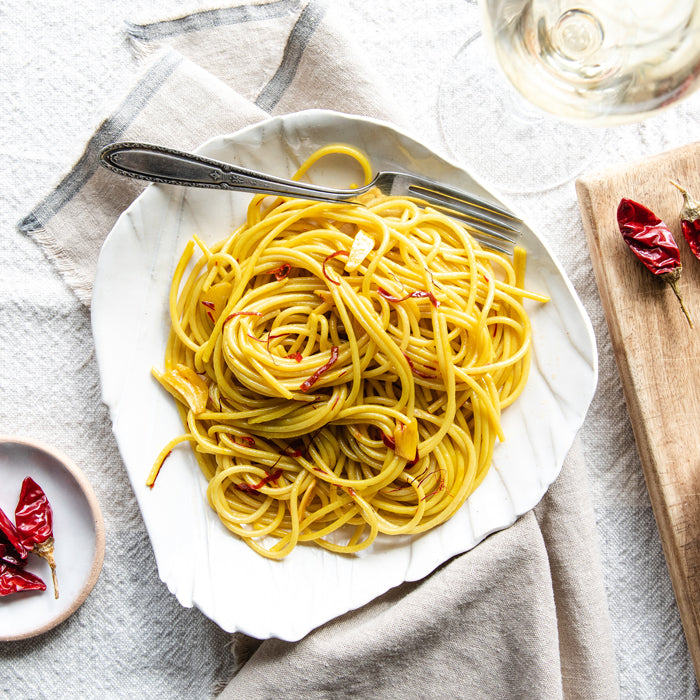 Saffron spaghetti with sliced garlic and red chili flakes on a white shell-shaped plate, fork and glass on light linen